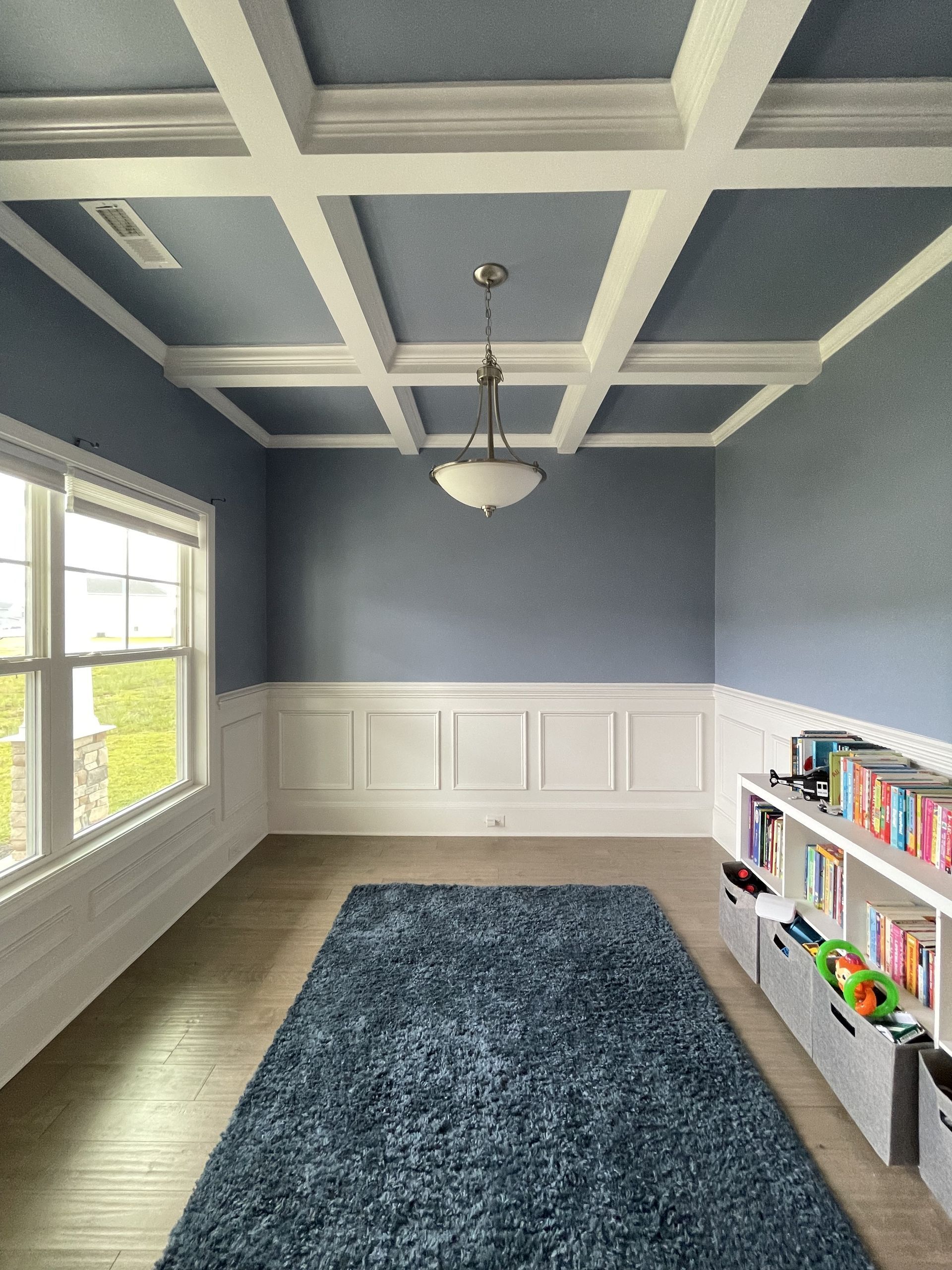 Empty bright room with blue walls, white coffered ceiling, large window, rug, and bookshelf.