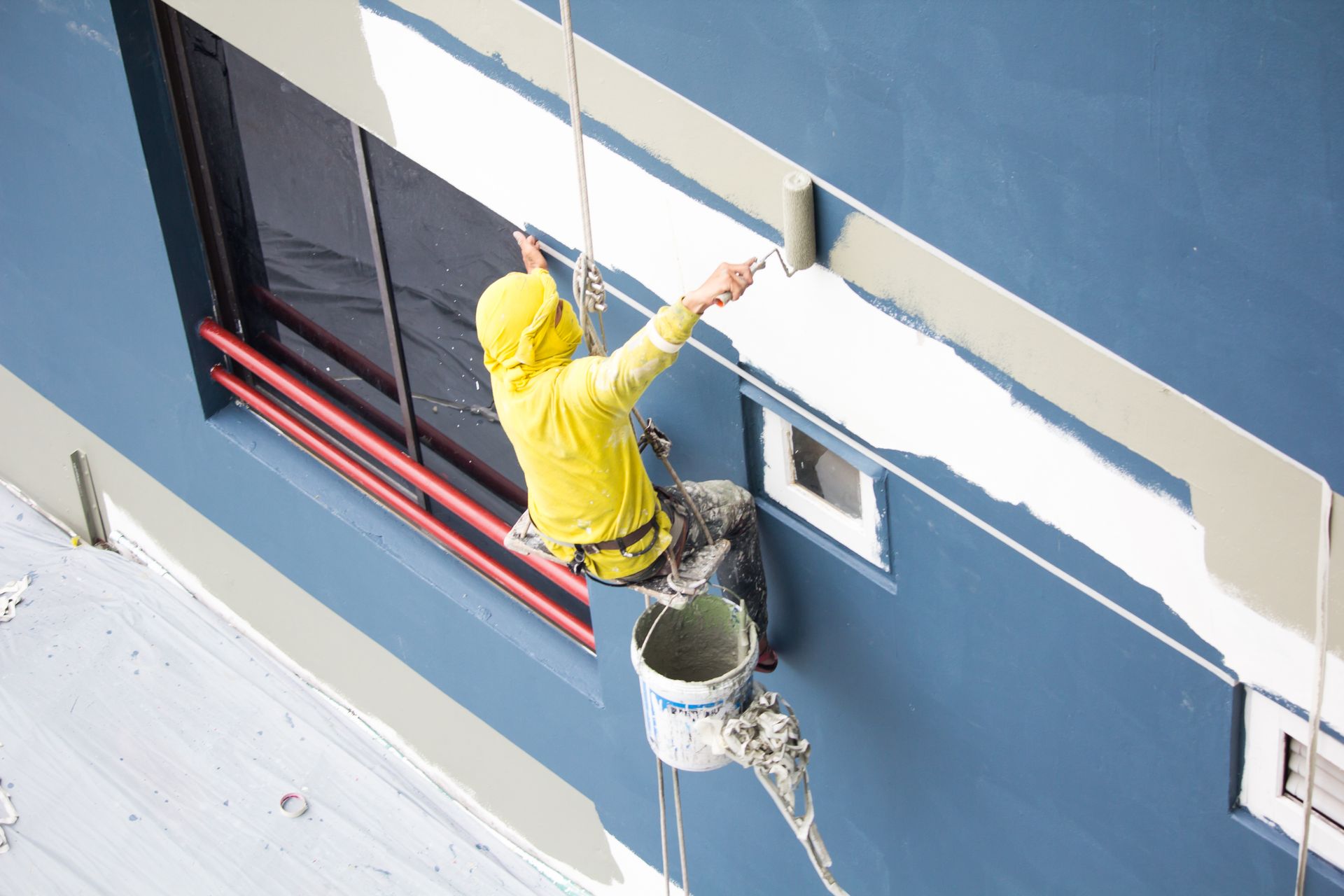 A worker in a yellow shirt hangs from ropes, using a roller to paint a white stripe on a blue building wall.