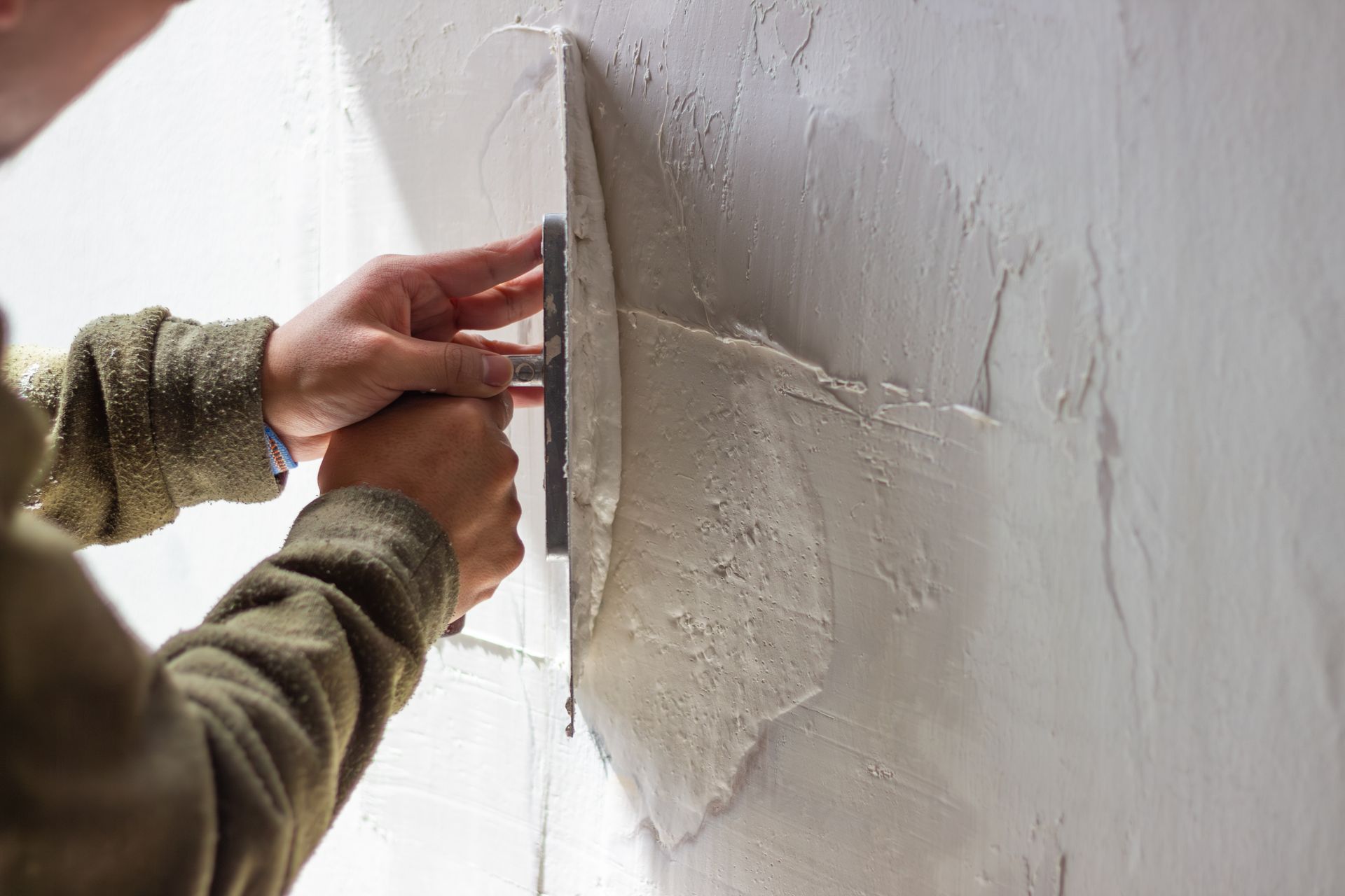 A person in a green long-sleeved shirt uses a metal trowel to spread gray plaster on a white wall.