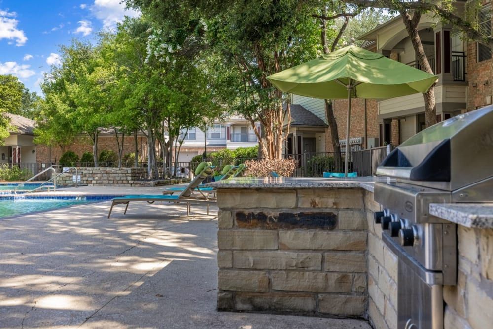 Community pool area with lounge chairs and a large green umbrella, surrounded by lush trees and residential buildings. A built-in outdoor grill is in the foreground at Brooks on Preston in Plano, TX.
