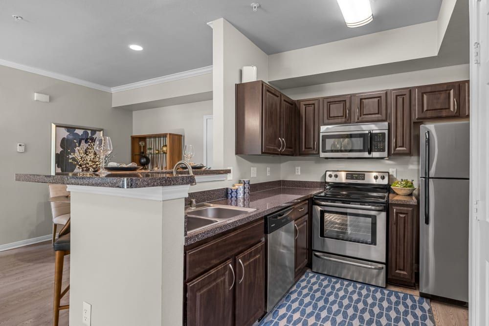 Modern kitchen interior with dark wooden cabinets, granite countertops, and stainless steel appliances. A breakfast bar with bar stools is visible, accented with a patterned blue rug on the floor. Decorative items such as a wine glass set and potted plants are arranged for an inviting atmosphere at Brooks on Preston in Plano, TX.