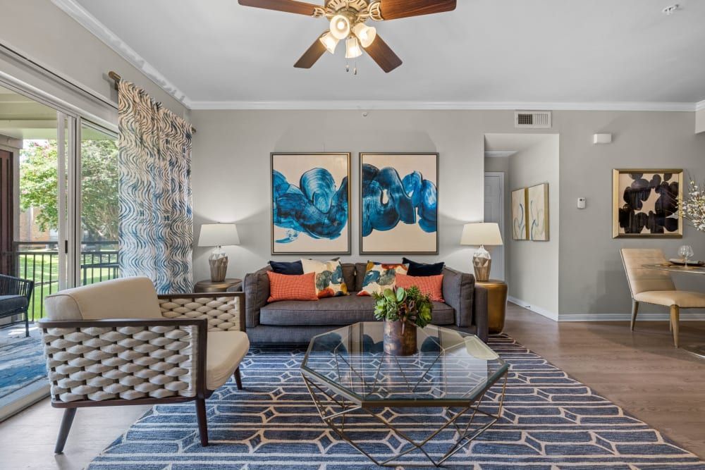 Modern living room interior with a geometric blue and white rug, a mid-century style tan armchair, and a dark gray sofa adorned with colorful pillows. The room features abstract blue artwork on the walls, a glass-top coffee table, and a ceiling fan. Natural light filters in through sliding glass doors that open to a balcony. To the right is a dining area with a wooden table and upholstered chairs at Brooks on Preston in Plano, TX.