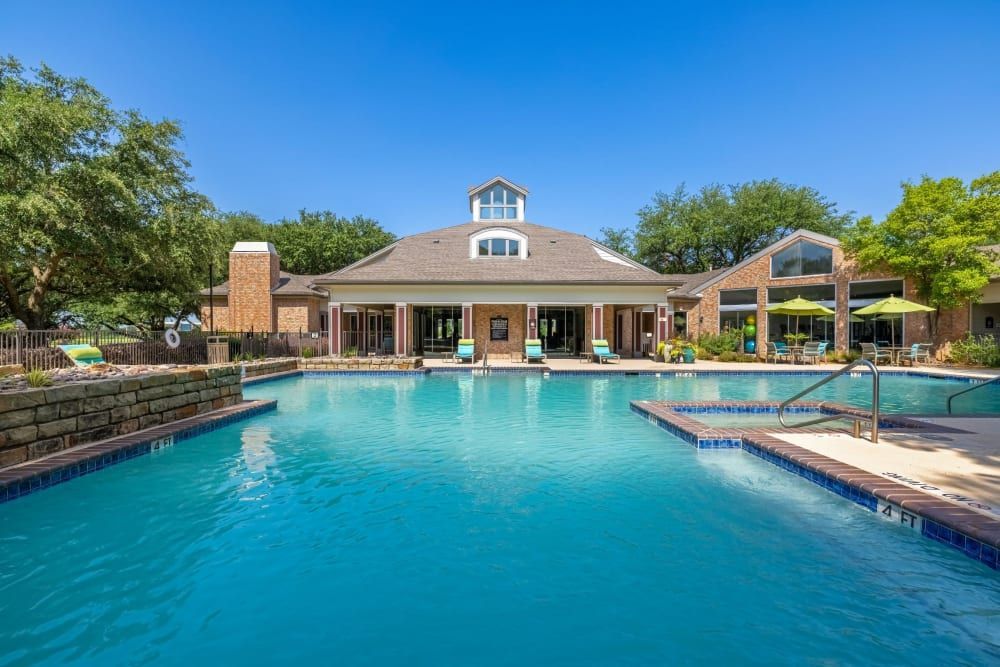 A spacious outdoor swimming pool with crystal clear water, surrounded by a well-maintained pool deck and brick fencing. A grand residential building with a cupola and large windows overlooks the pool, featuring loungers and bright yellow umbrellas that provide shade. The scene conveys a relaxed and luxurious backyard setting on a sunny day with a cloudless blue sky at Brooks on Preston in Plano, TX.