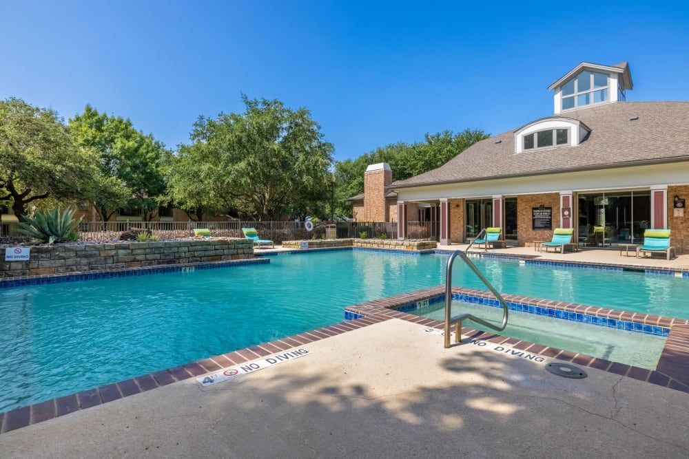 Spacious outdoor swimming pool area with clear blue water, surrounded by a neat tile deck and a well-maintained yard with lush green trees. The scene includes a two-story house with brick facade and large windows, poolside furniture for relaxation, and clear blue sky above at Brooks on Preston in Plano, TX.