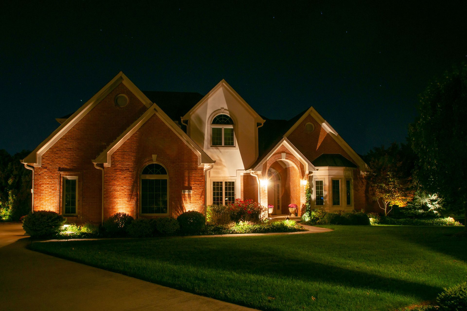 Lit-up brick house at night; landscape lighting illuminates lawn and exterior; dark blue sky background.