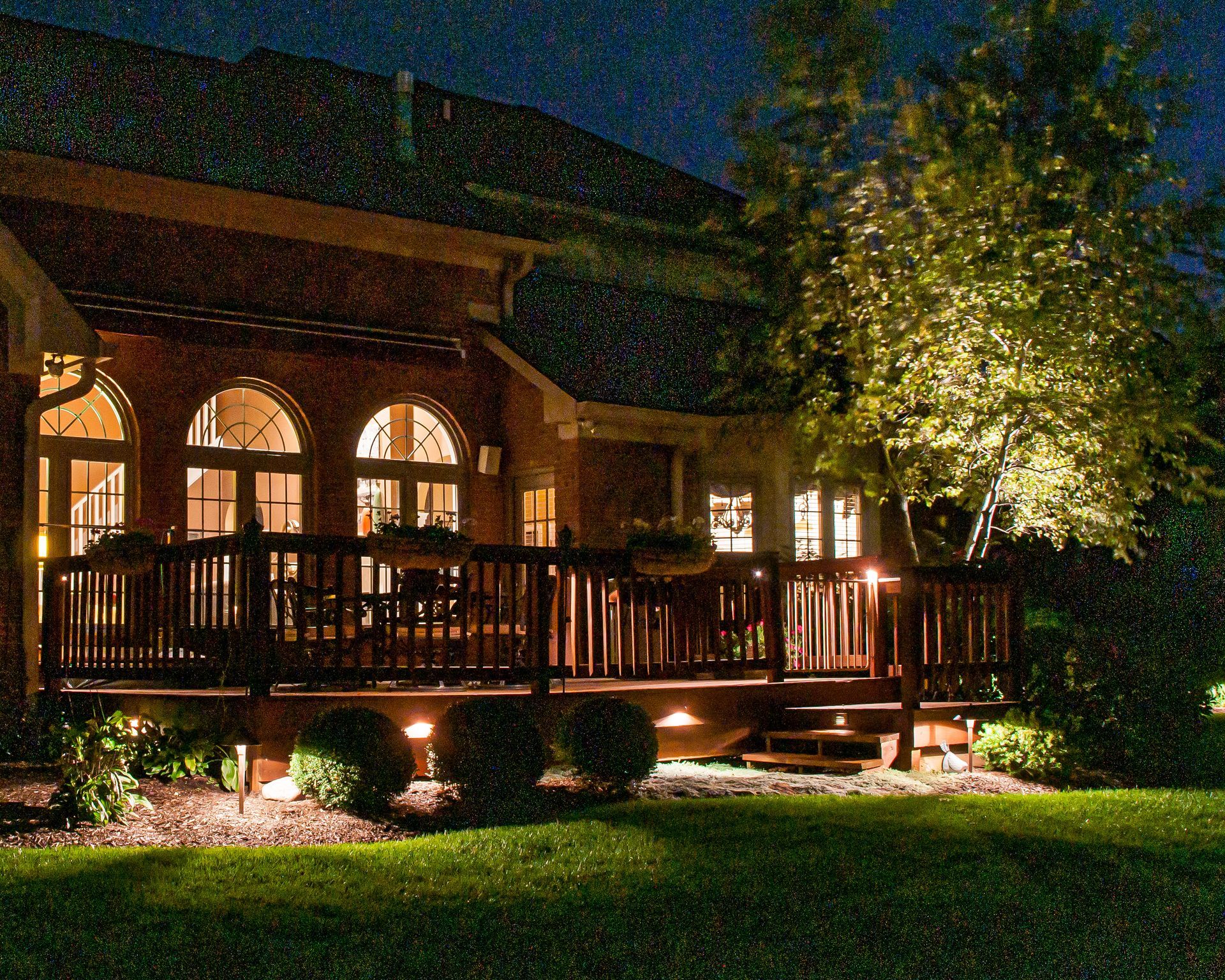 Nighttime view of a wooden deck with landscape lighting; lit house in the background.