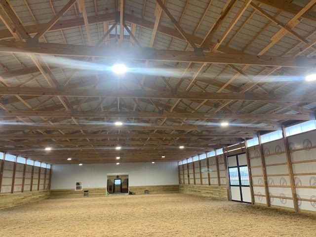 Indoor equestrian arena with sand footing, wood beams, and bright lights.