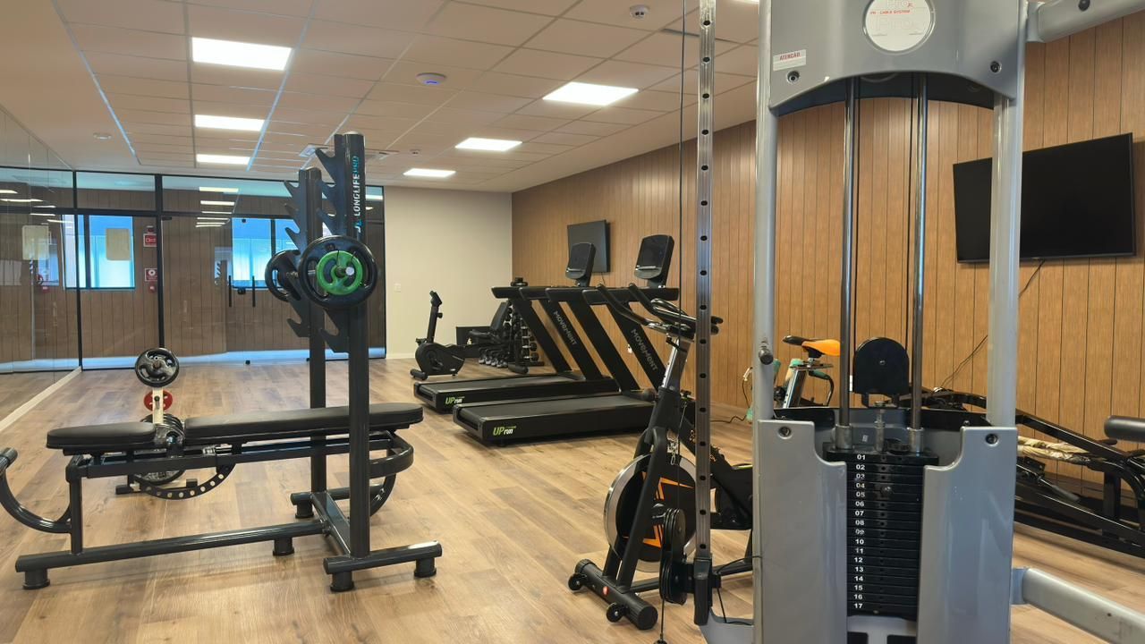Interior de ginásio com equipamentos de ginástica, incluindo esteiras e máquinas de musculação, em frente a painéis de madeira.