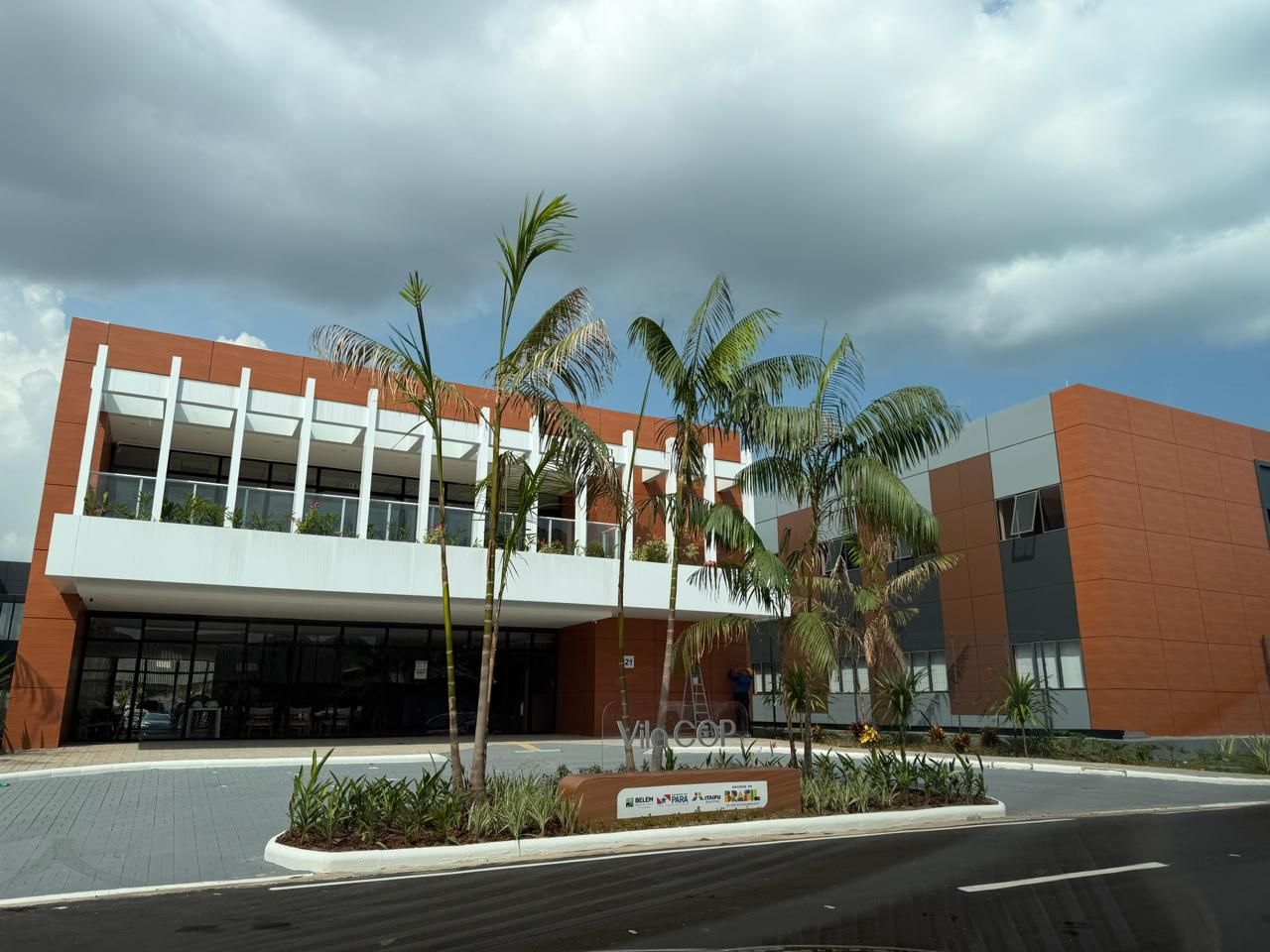 Prédio moderno de tijolos com detalhes em branco, palmeiras e céu nublado.