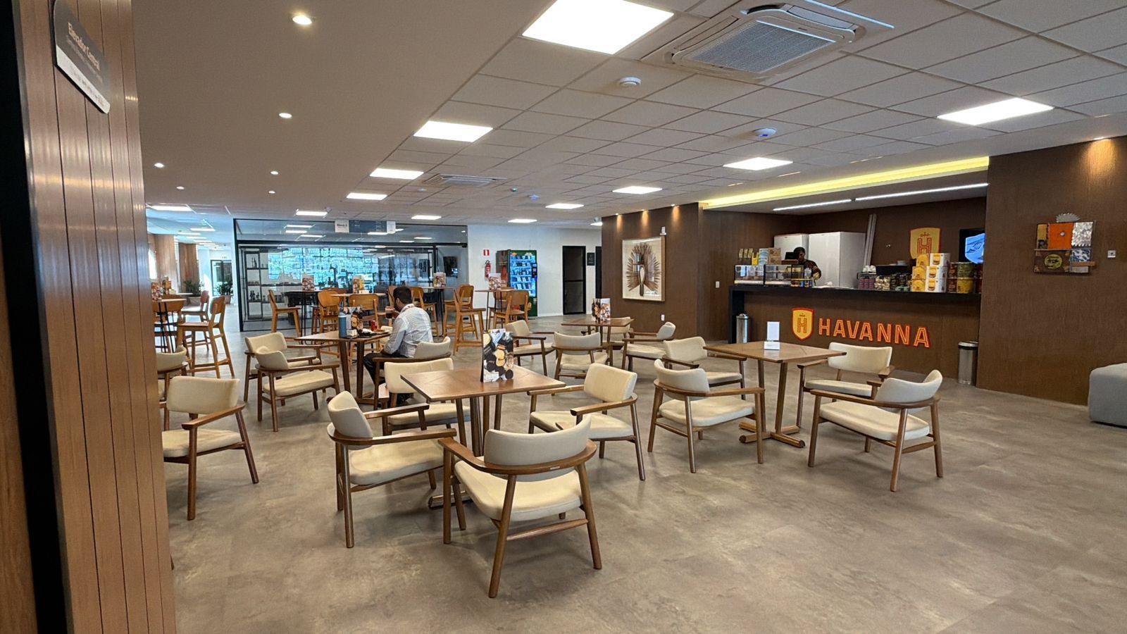 Interior de um café com mesas, cadeiras e balcão. Pessoas sentadas. O café possui detalhes em madeira e iluminação clara.