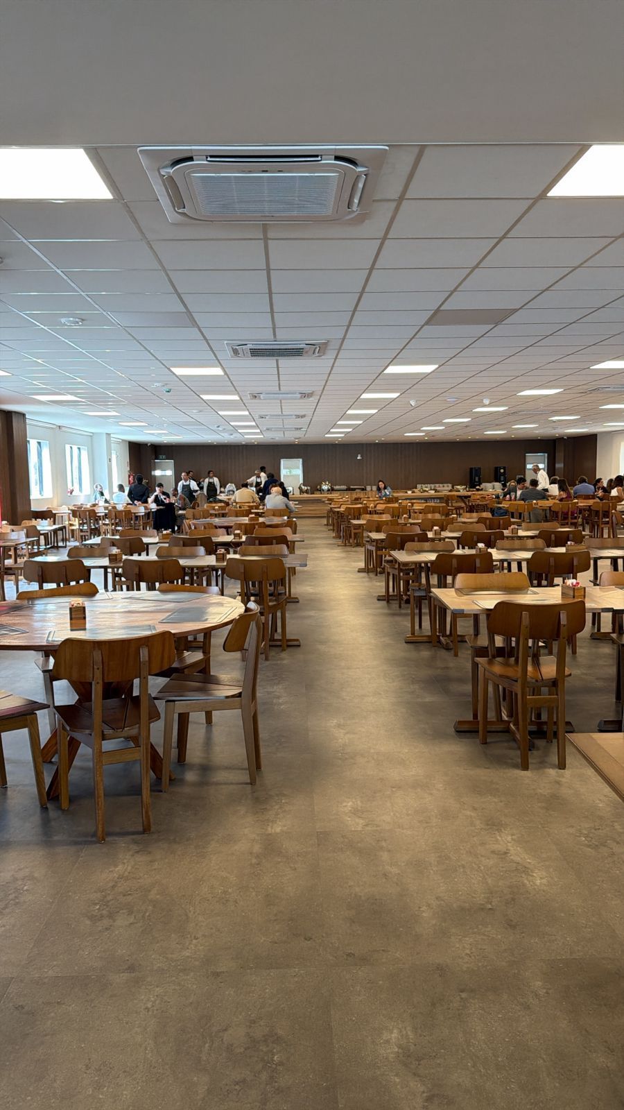 Um refeitório grande e vazio, com muitas mesas e cadeiras de madeira, e um longo balcão de atendimento no fundo.