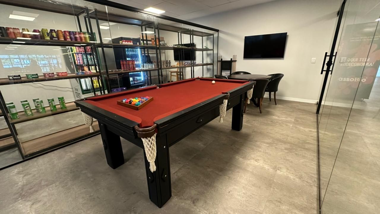 Uma sala de jogos com mesa de bilhar, lanches e assentos. Mesa de bilhar vermelha, pés pretos e prateleiras.