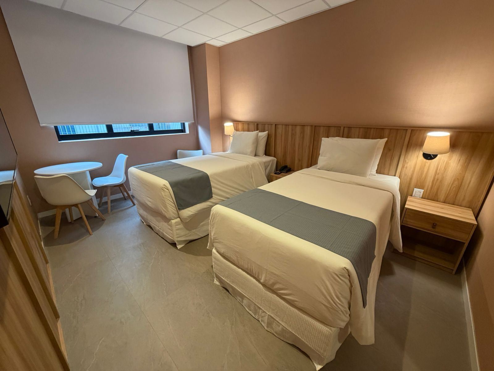 Quarto de hotel com duas camas de solteiro, mesas de cabeceira, uma mesinha de centro e uma janela com persiana.