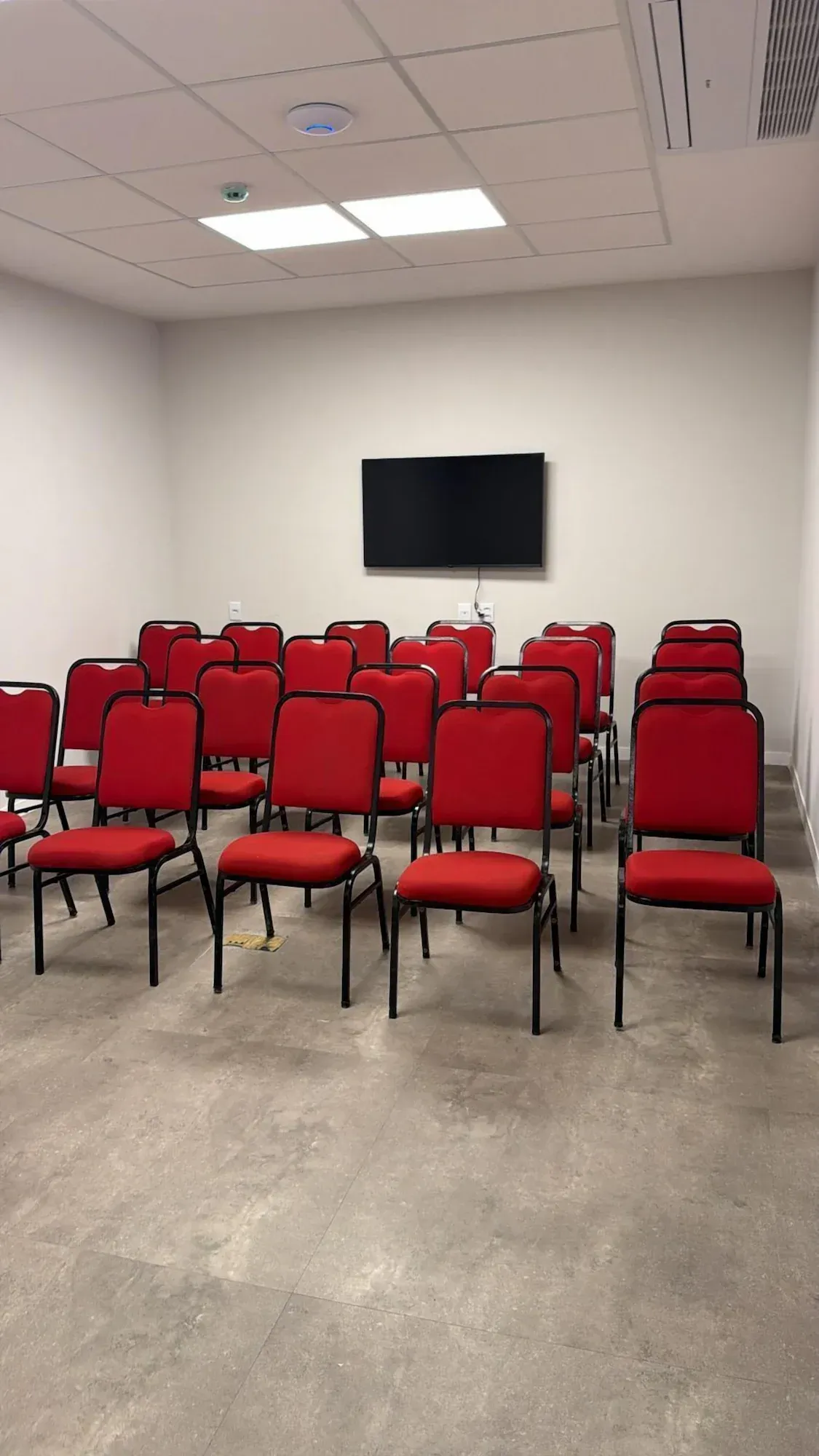 Cadeiras vermelhas dispostas em fileiras estão voltadas para uma tela preta em uma sala de paredes brancas.