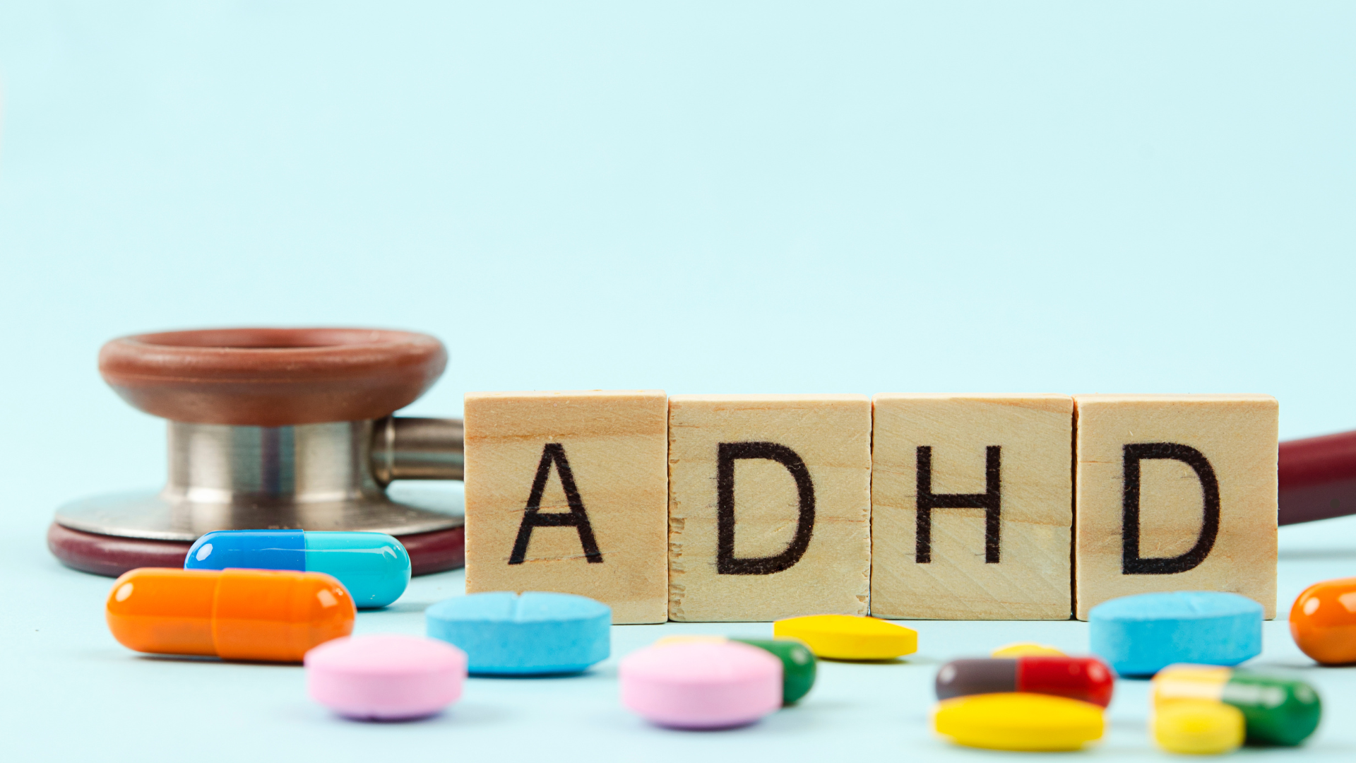 Wooden blocks spelling “ADHD” beside scattered colorful medication pills and a stethoscope, represen