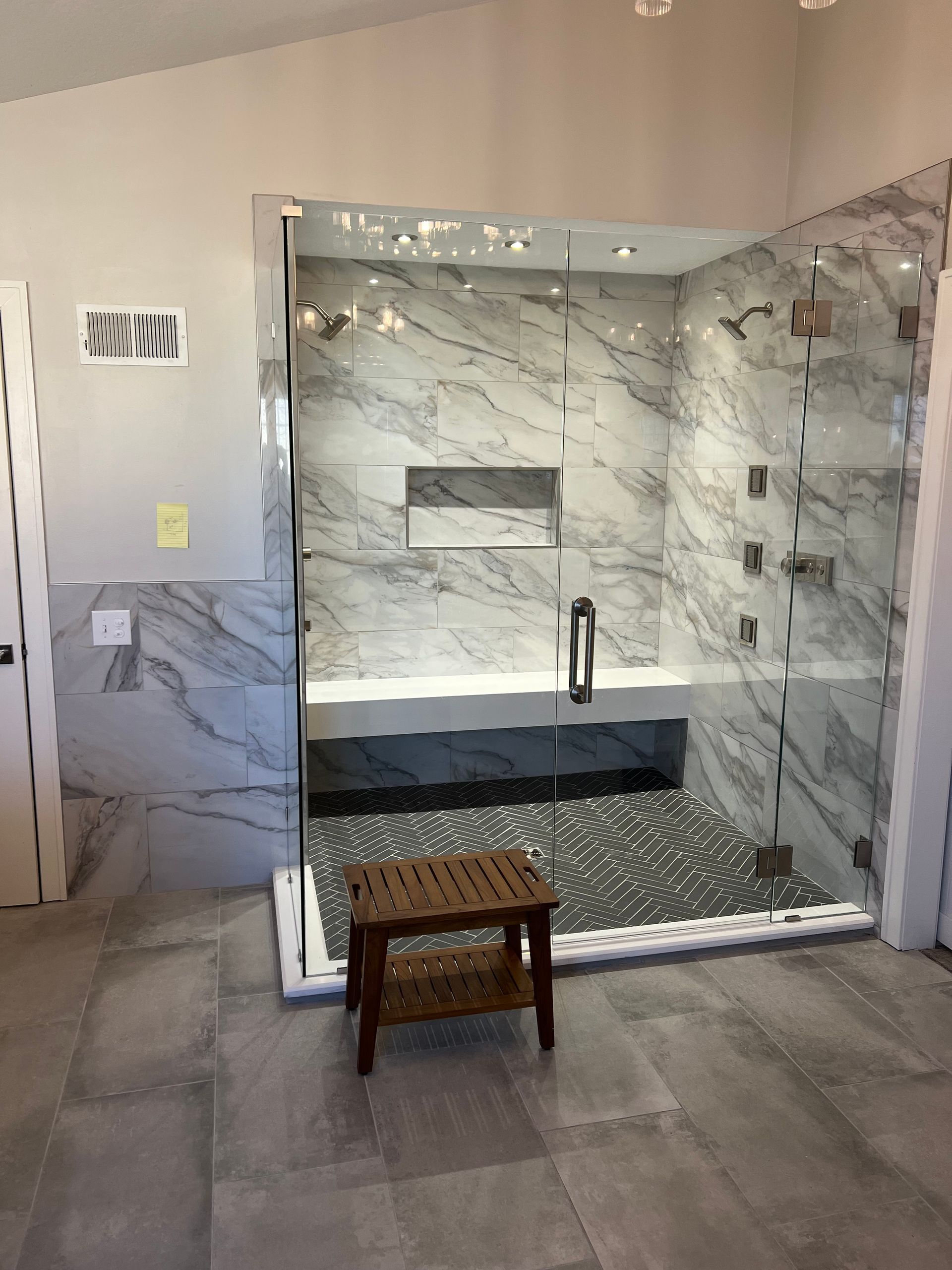 Glass-enclosed shower with marble-look tile, bench, and wooden stool on stone floor.