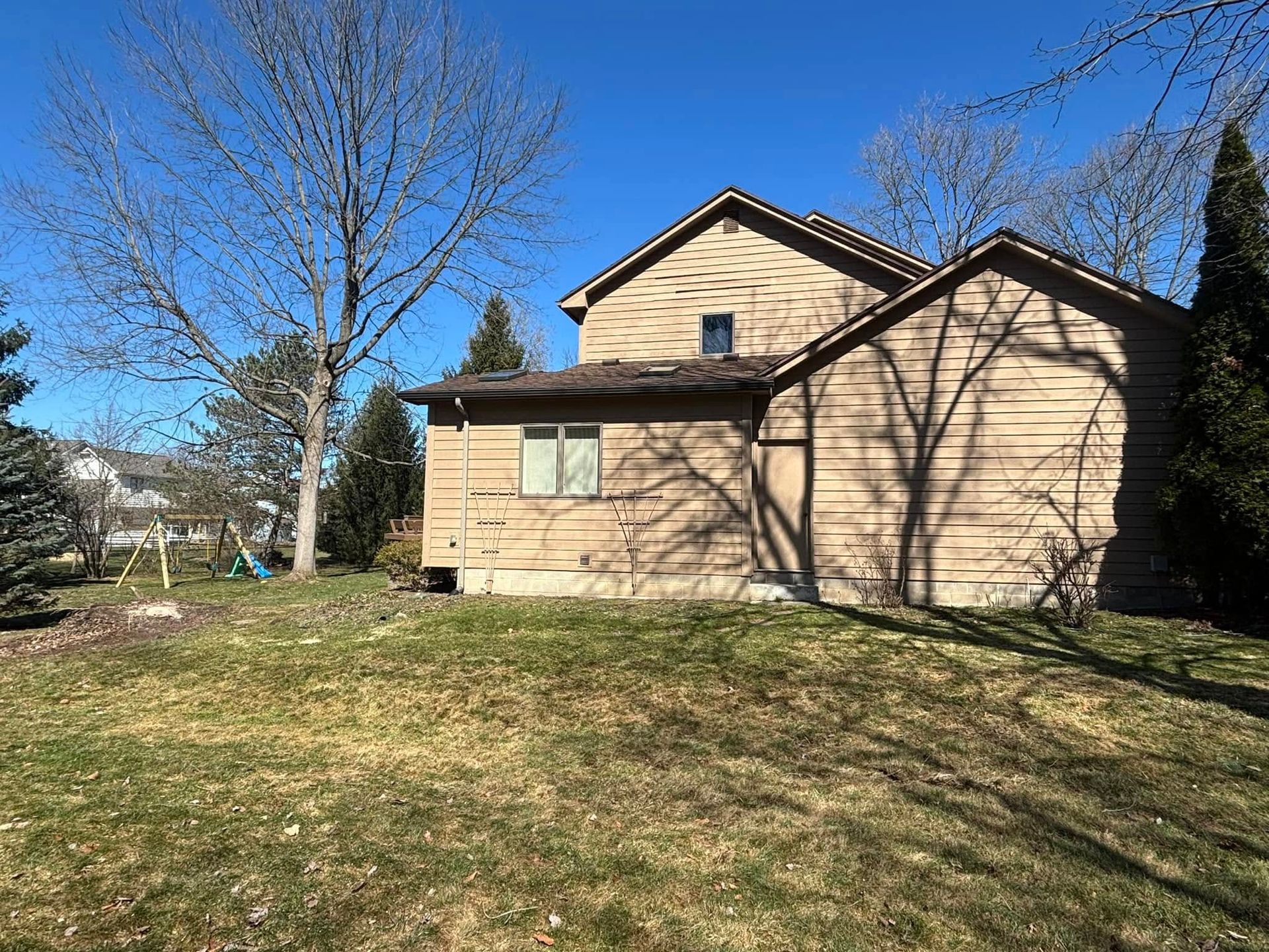 A tan, multi-level house with brown siding stands in a grassy yard under a bright blue sky, surrounded by tall trees.