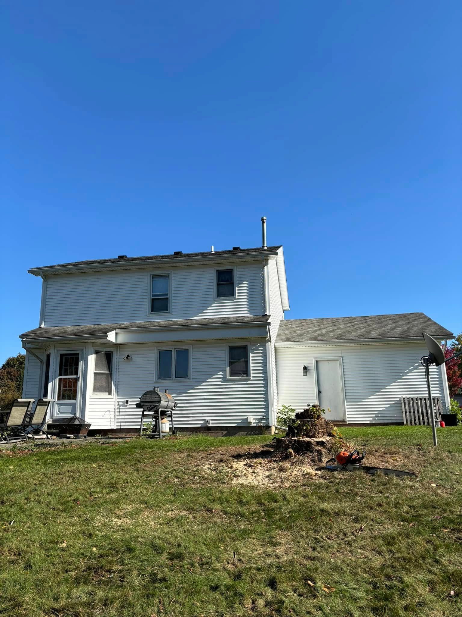 A white two-story house with a single-story wing, a grill on the lawn, and a pile of yard debris under a clear blue sky.
