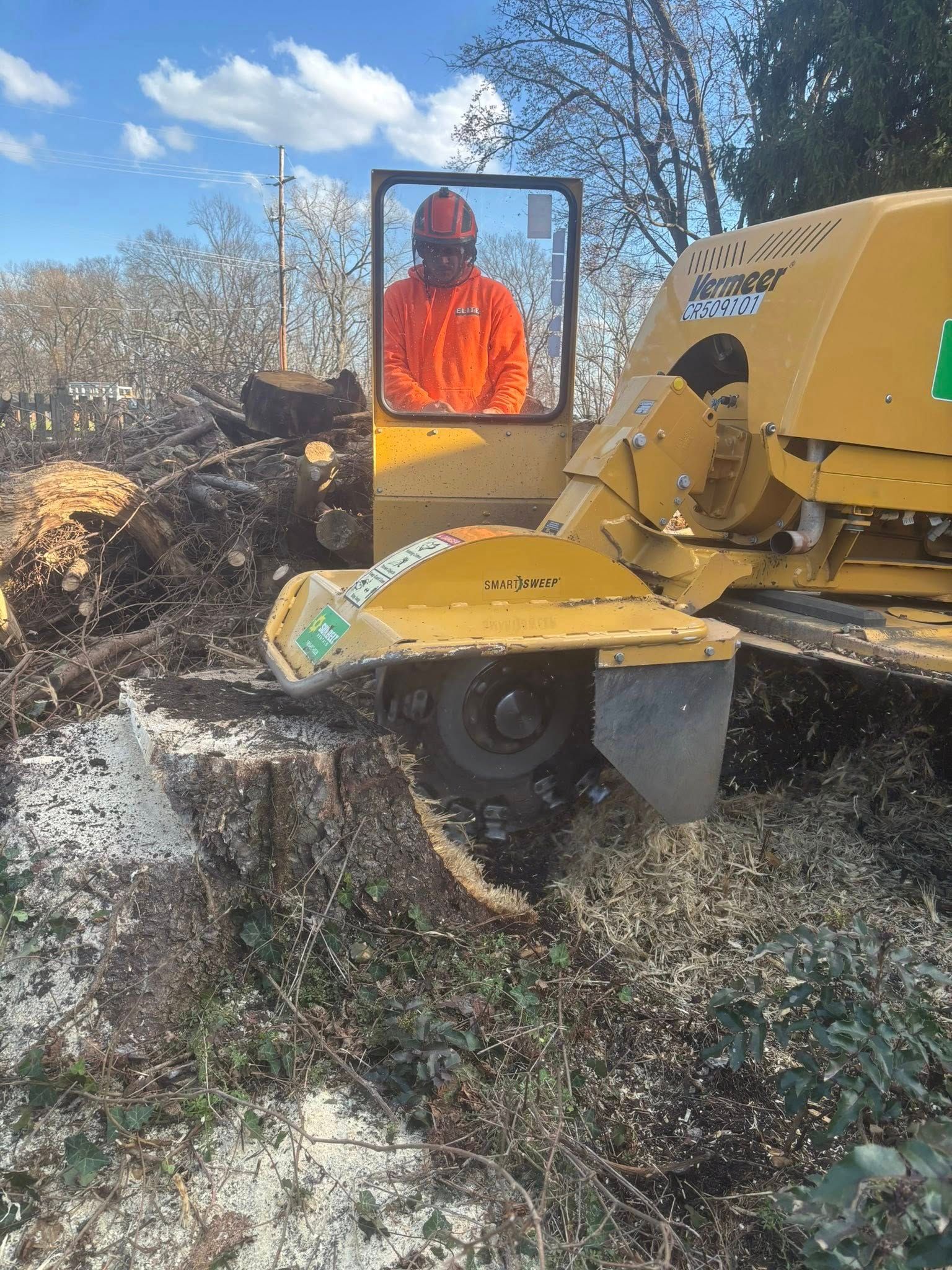 A person wearing high-visibility orange gear operates a yellow stump grinder to clear wood debris outdoors.
