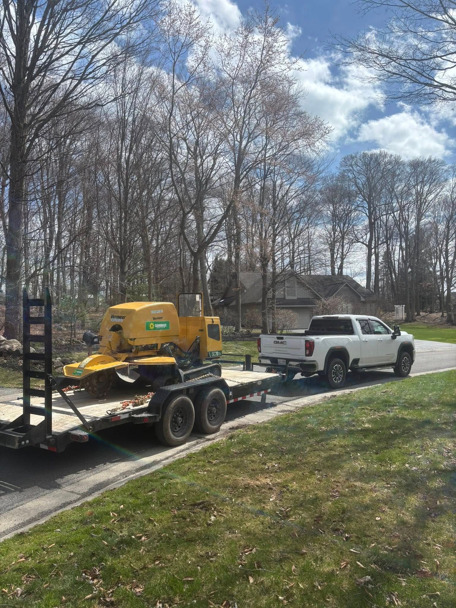 A white pickup truck hauls a yellow wood chipper on a trailer along a driveway lined with trees.