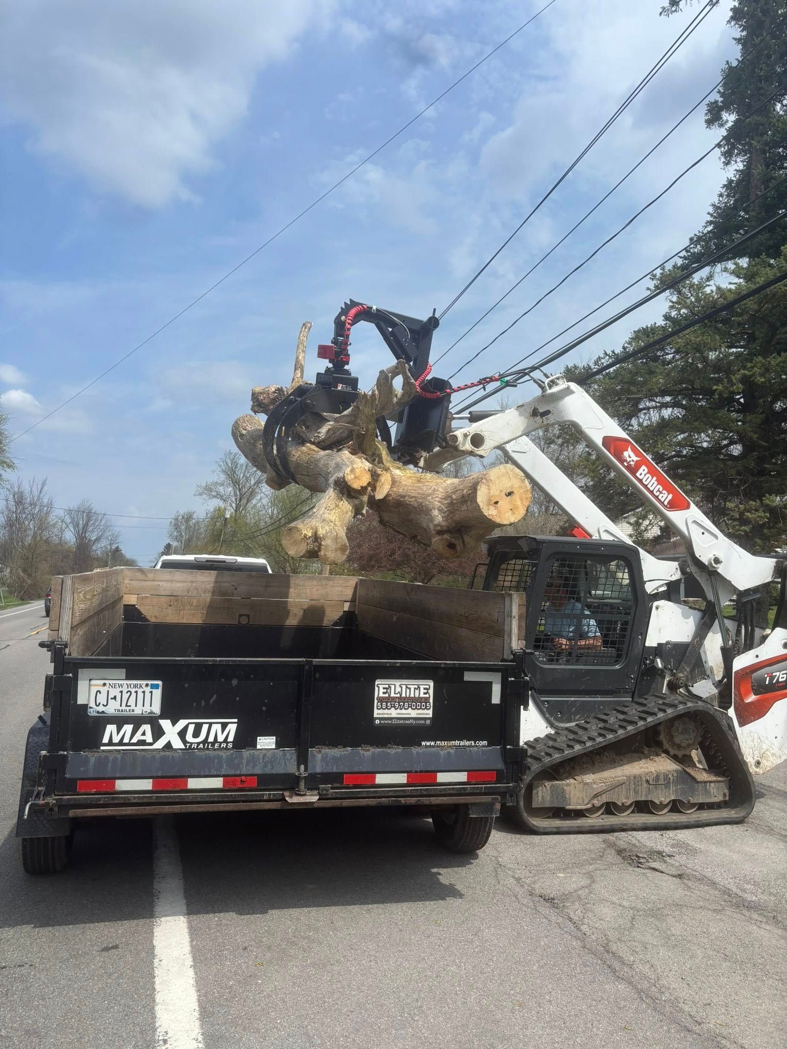 A skid steer loader lifts a large section of a tree trunk to load it into the bed of a black dump trailer on a road.