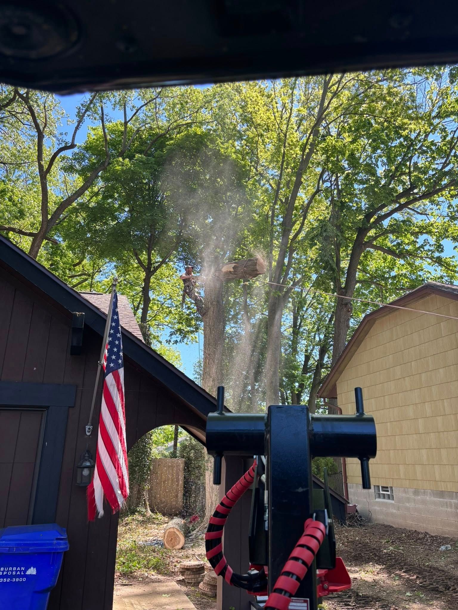 An American flag hangs on a dark building next to machinery during outdoor tree trimming work.