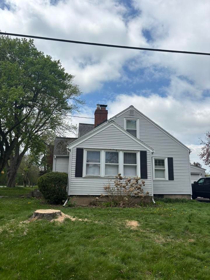 A light gray, single-story house with black shutters, a brick chimney, and a freshly cut tree stump in the front yard.
