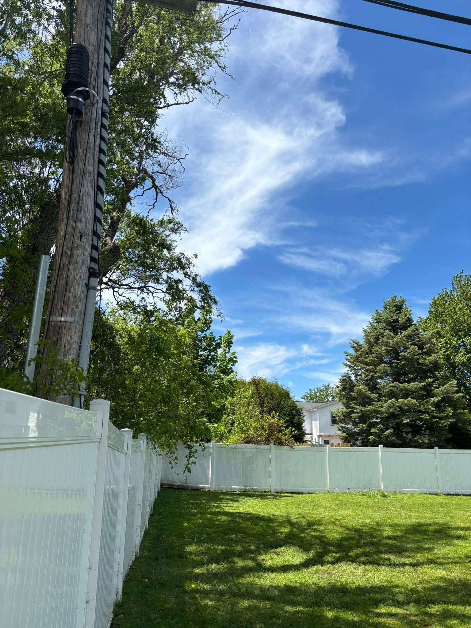 A white vinyl fence borders a green grassy backyard under a bright blue sky with wispy clouds and a utility pole.