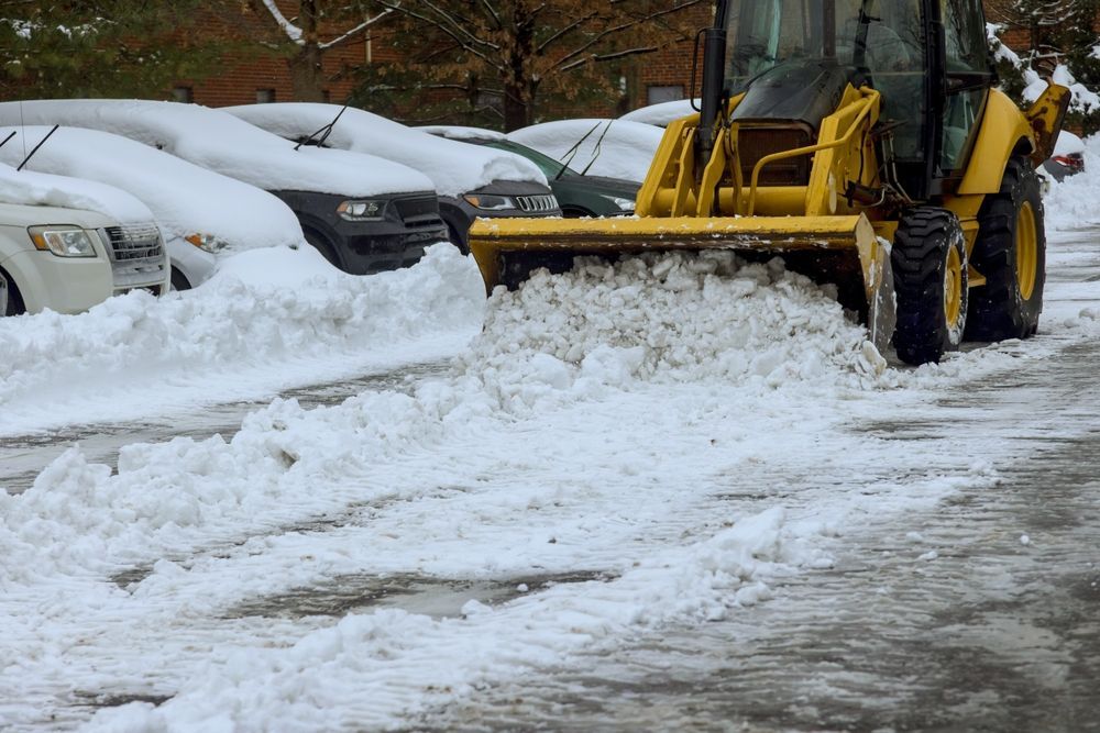 A yellow front-end loader clearing deep snow from a parking lot filled with snow-covered cars.
