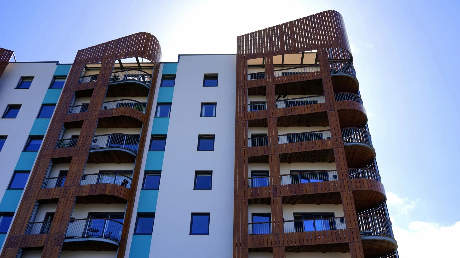 Multi-story apartment building with brown wood accents, turquoise details, and balconies against a blue sky.