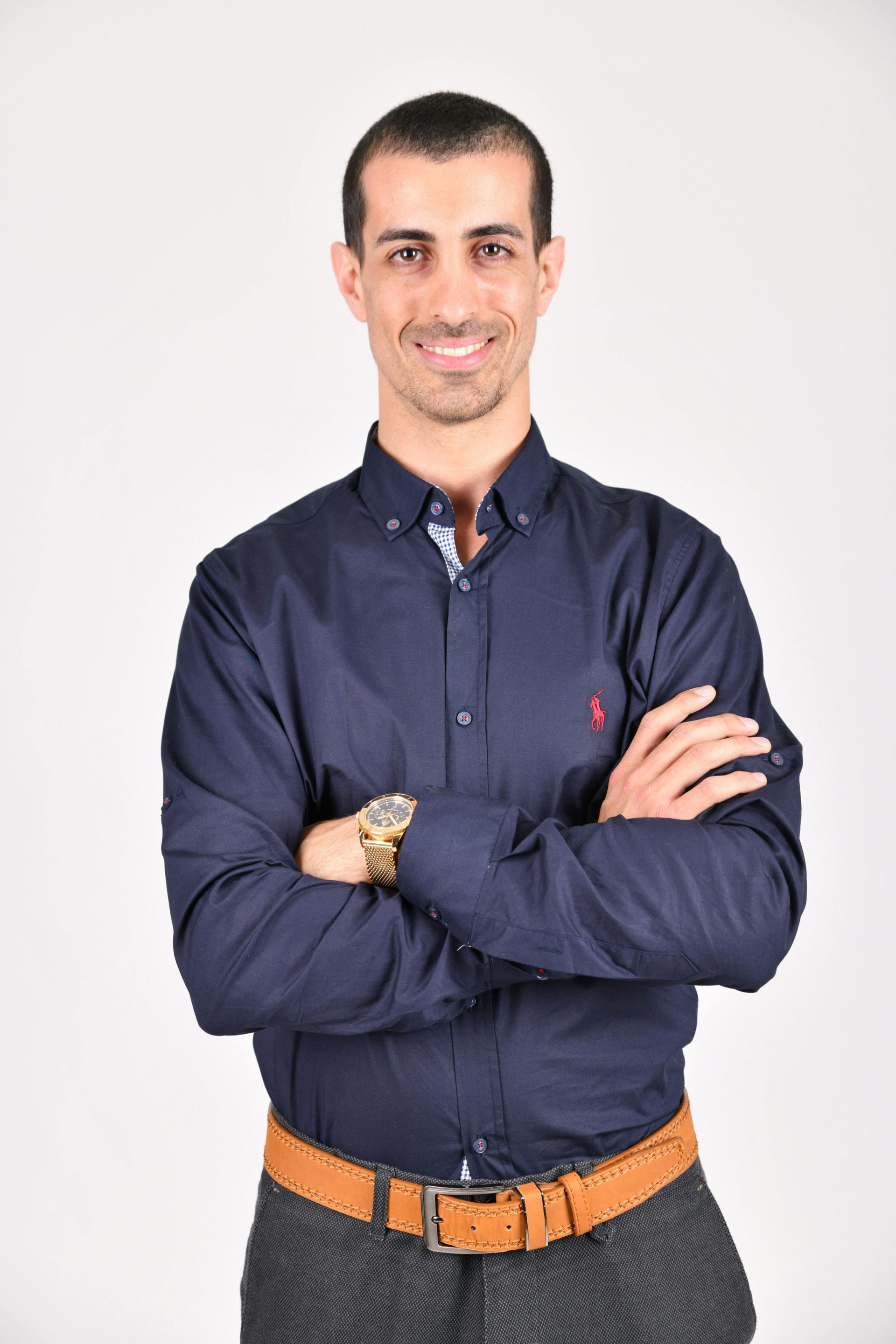 Man with arms crossed, wearing a blue shirt and grey pants, smiling at the camera.
