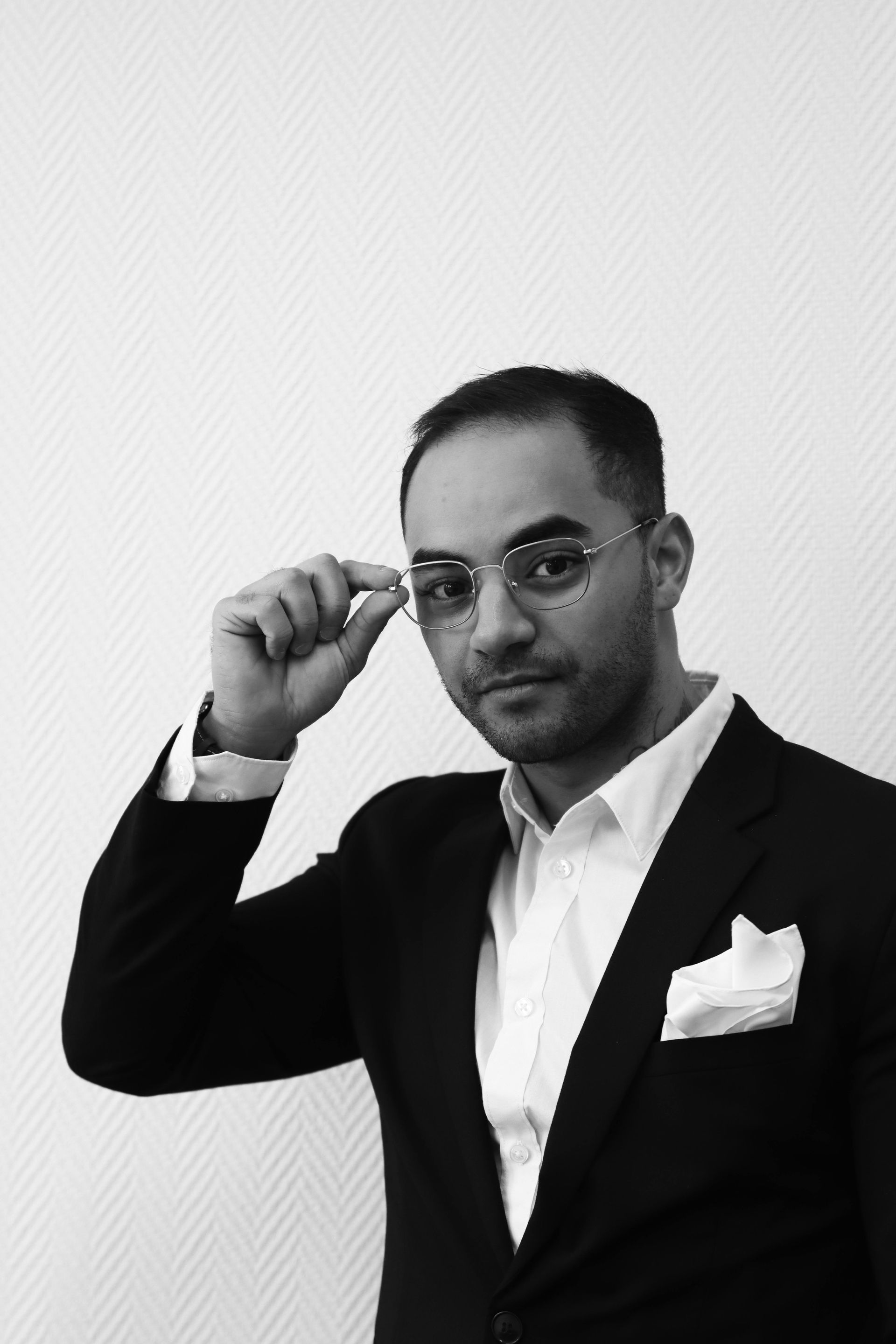 Man in a suit adjusts glasses, posing for the camera with a neutral expression.