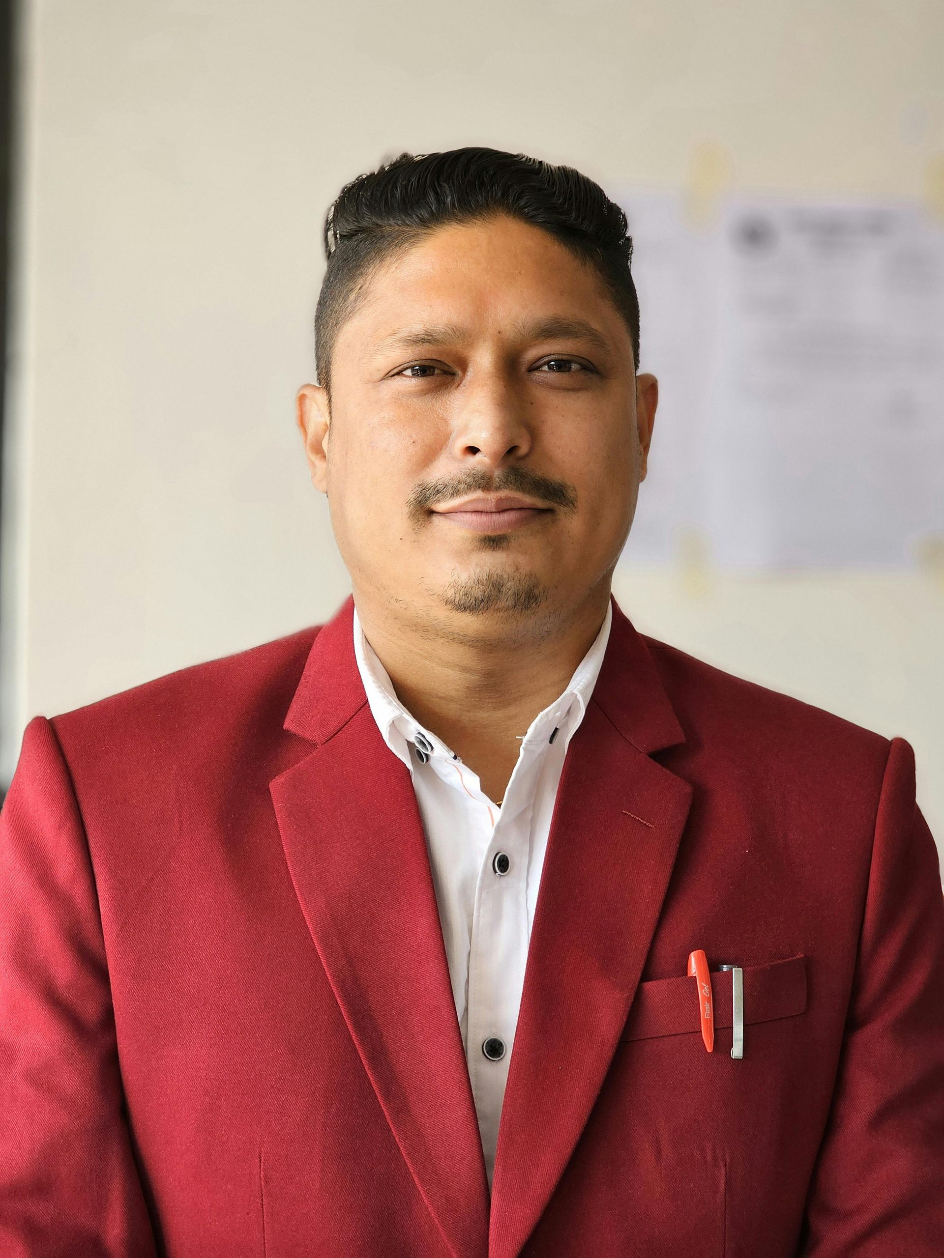 Man in red blazer and white shirt, smiling, pens in pocket.
