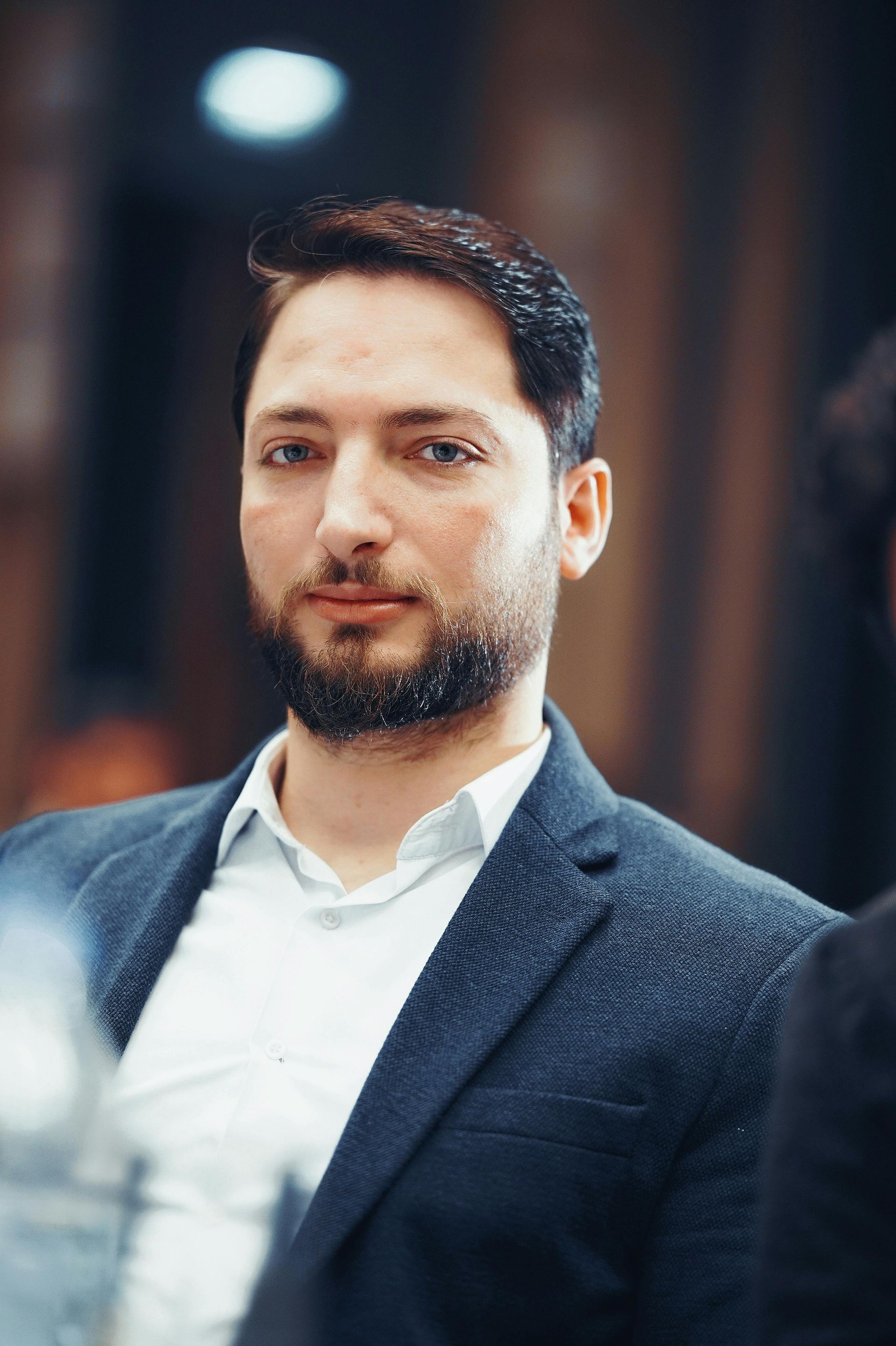 Man in a navy blazer and light blue shirt, looking toward the camera with a slight smile.