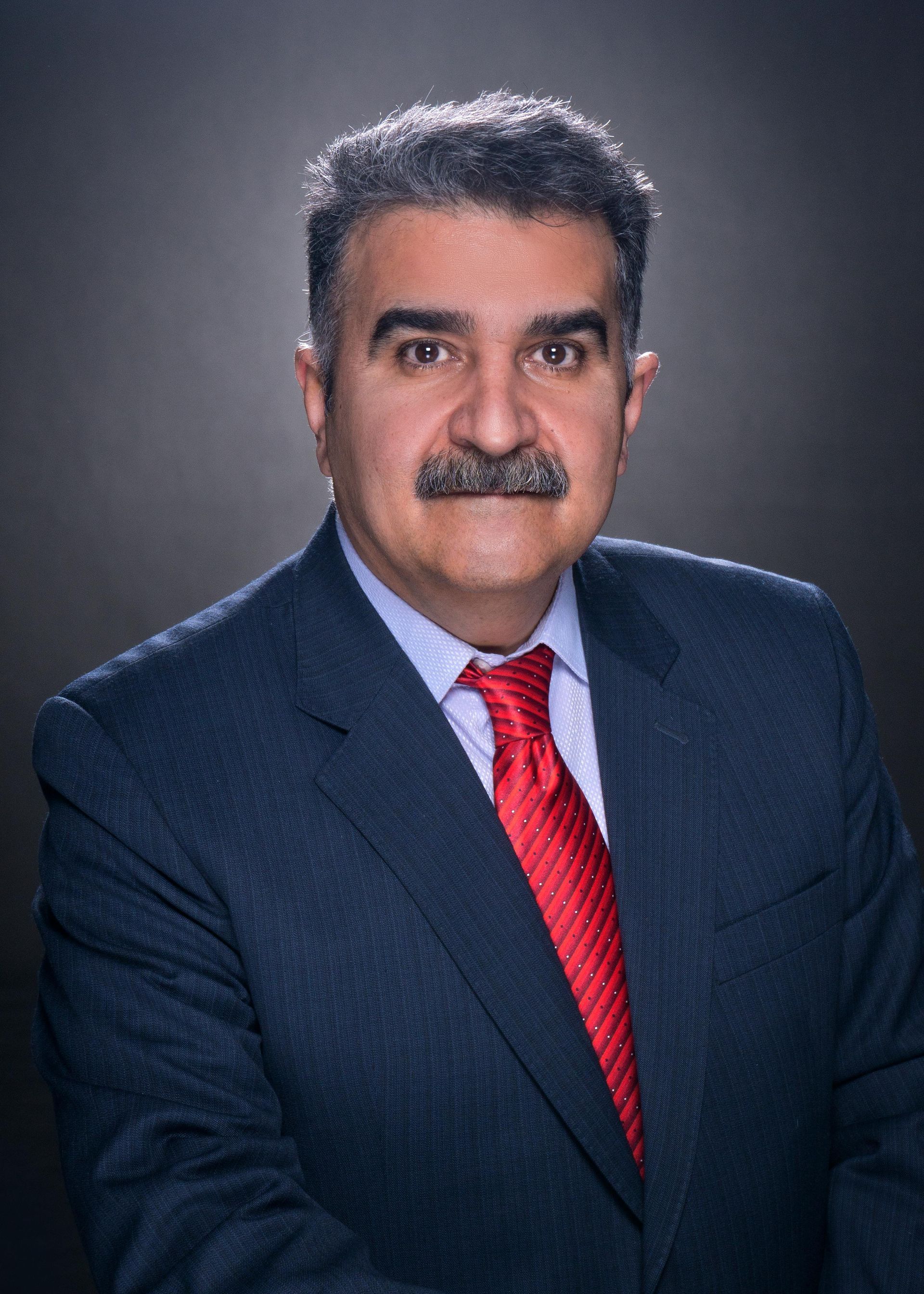 Man in suit and red tie, smiling with a mustache, against a dark gray background.