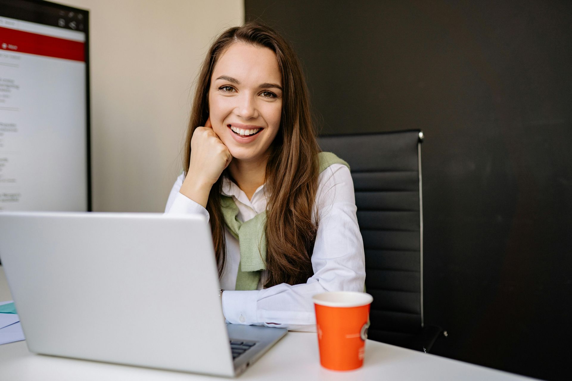 Woman smiles at a desk with a laptop and coffee cup; a dark wall in the background.