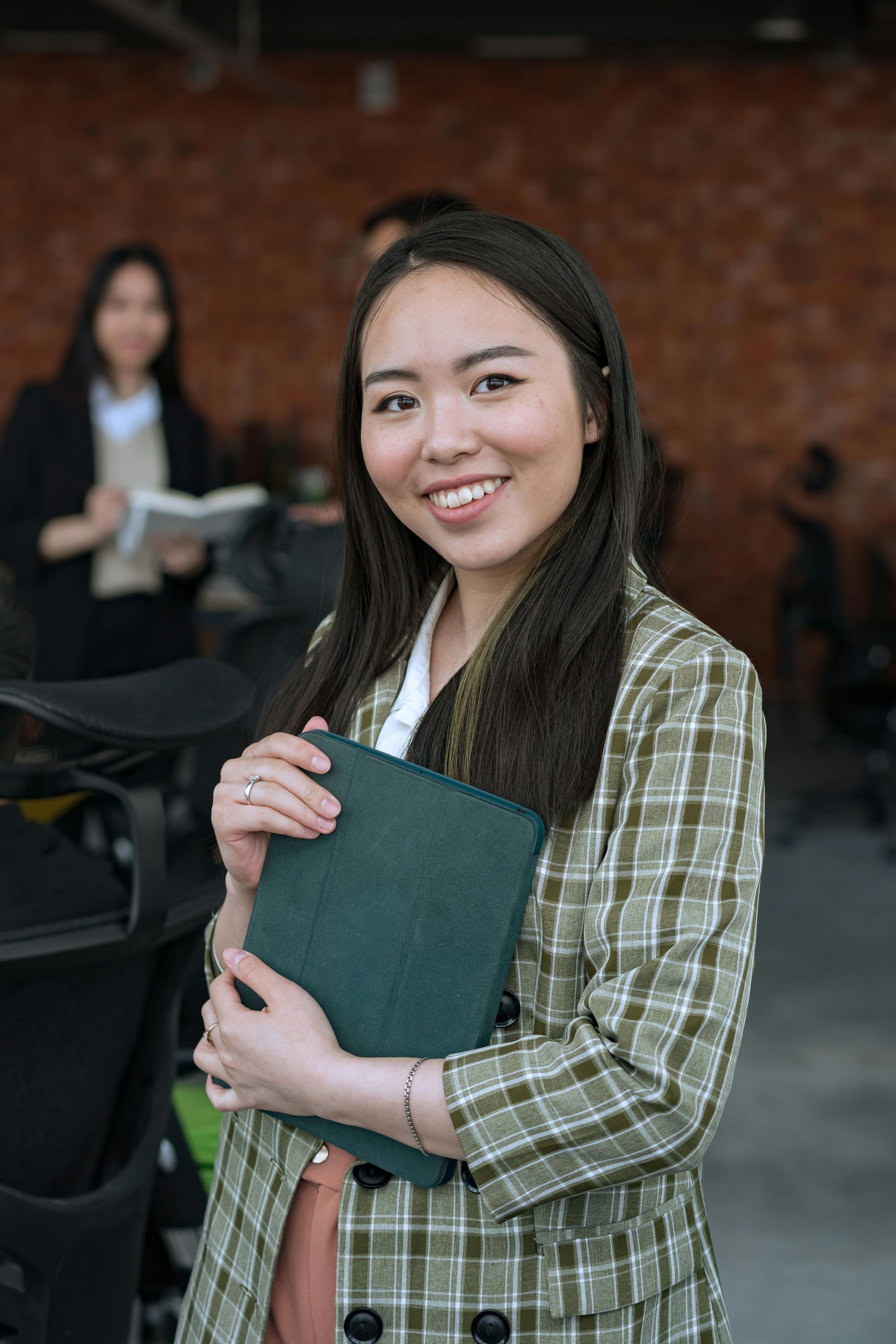 Woman with a green folder, wearing a plaid blazer, smiles in a room. Another person is in the background.