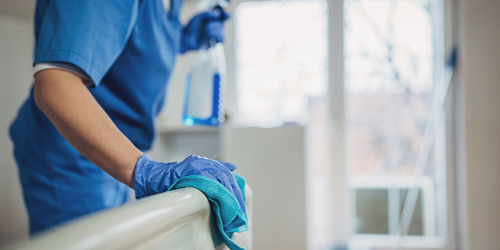 Person wearing blue scrubs and gloves sprays and wipes a surface with a cloth.