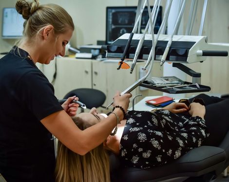 Dentist examining a patient in a dental chair. Blonde woman in black outfit, dental tools visible.