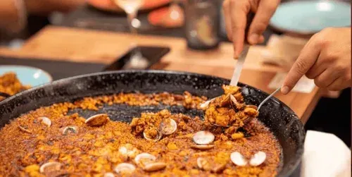 Hands serving a large pan of orange rice dish with mussels on a table