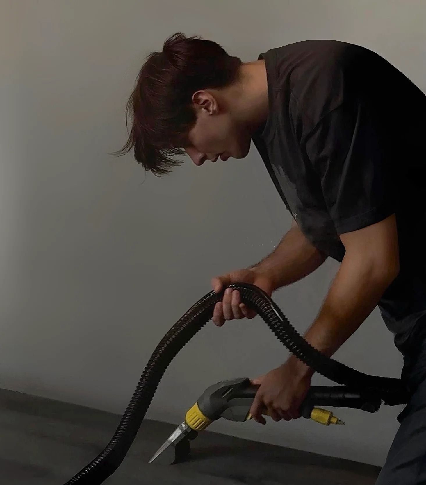 Person vacuuming a surface, holding hose and nozzle, wearing dark shirt, in a room.