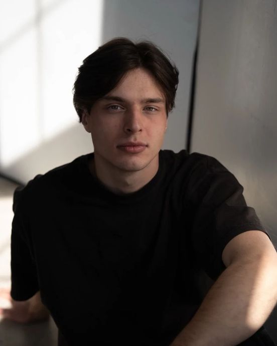 Man with dark hair and a black shirt looking directly at the camera in a shadowed setting.