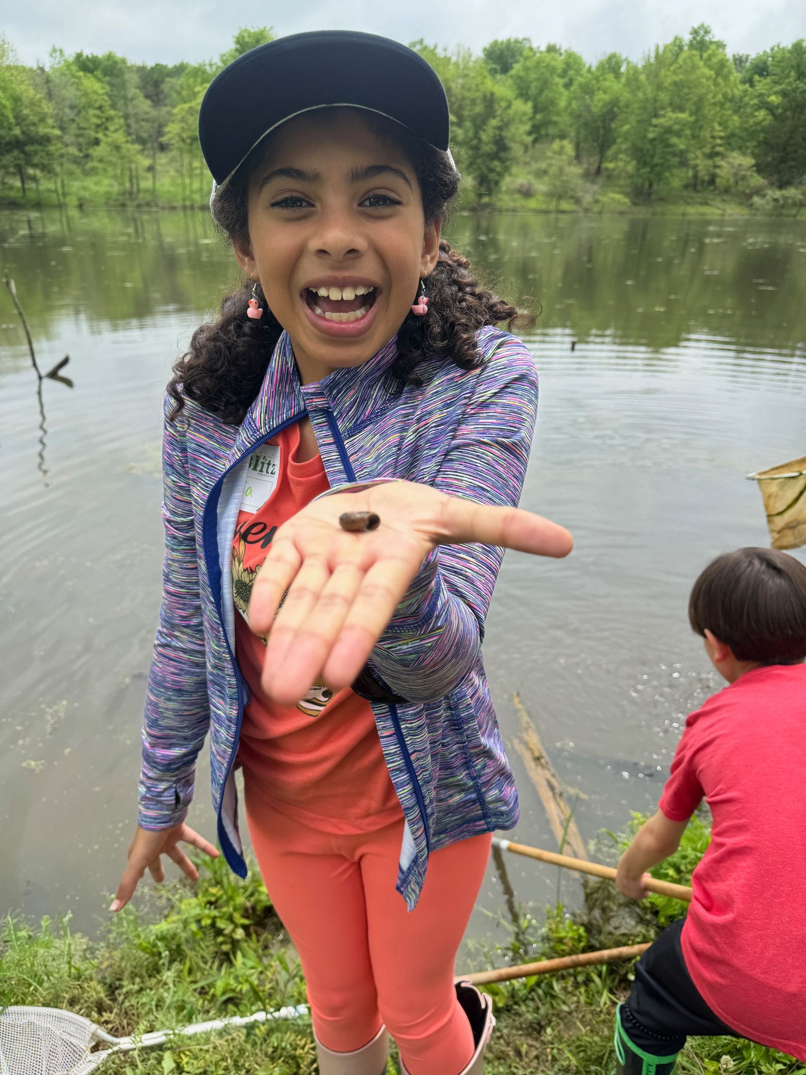 Smiling child in a cap shows a tiny frog near a pond, with another child crouching by the water.
