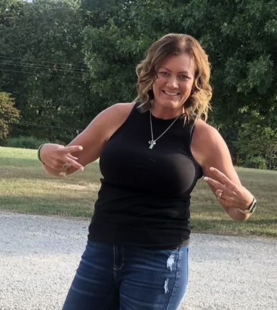 A woman in a black tank top and blue jeans is giving a peace sign.