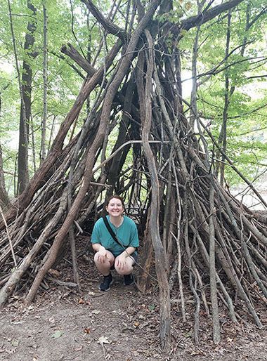 A woman is kneeling in front of a wooden structure in the woods.