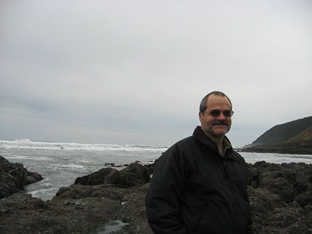 A man in a black jacket is standing on a rocky beach near the ocean.