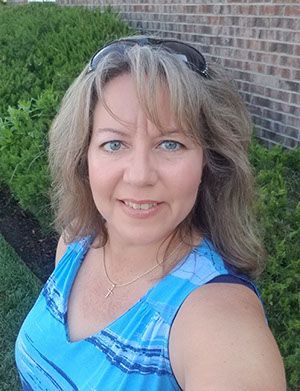 A woman in a blue tank top is taking a selfie in front of a bush.