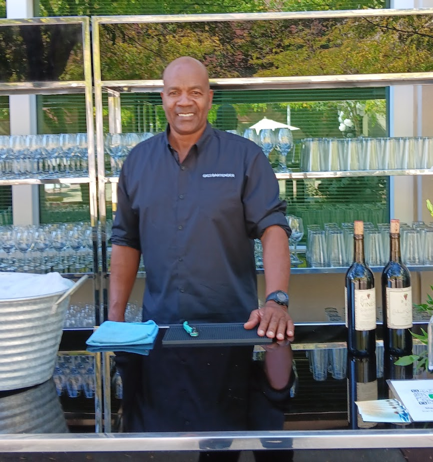 bartender smiles, facing camera. Bottles of wine sit beside him. 