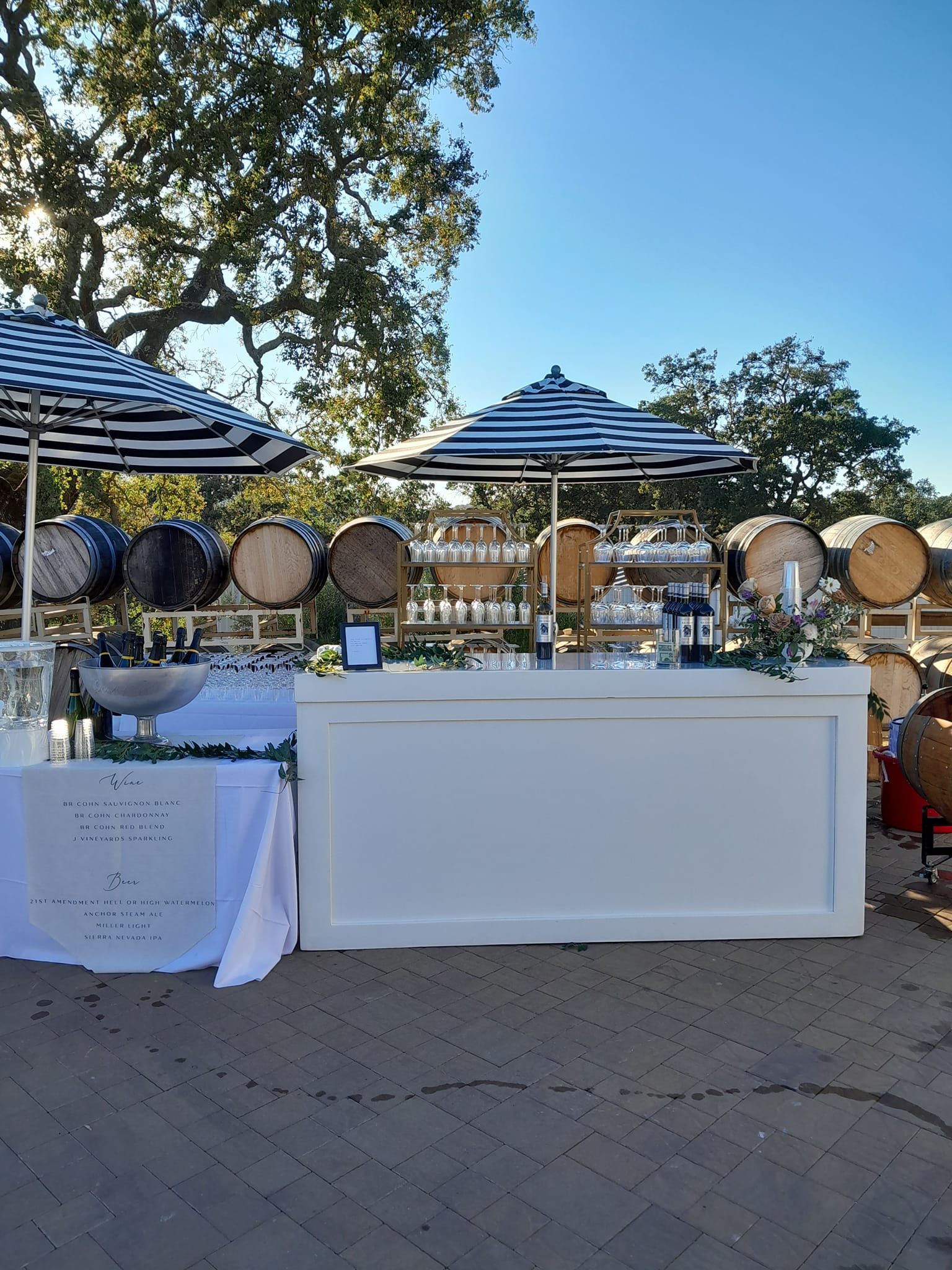 Outdoor white bar with striped umbrellas, barrels, and drinks. Sunny day.
