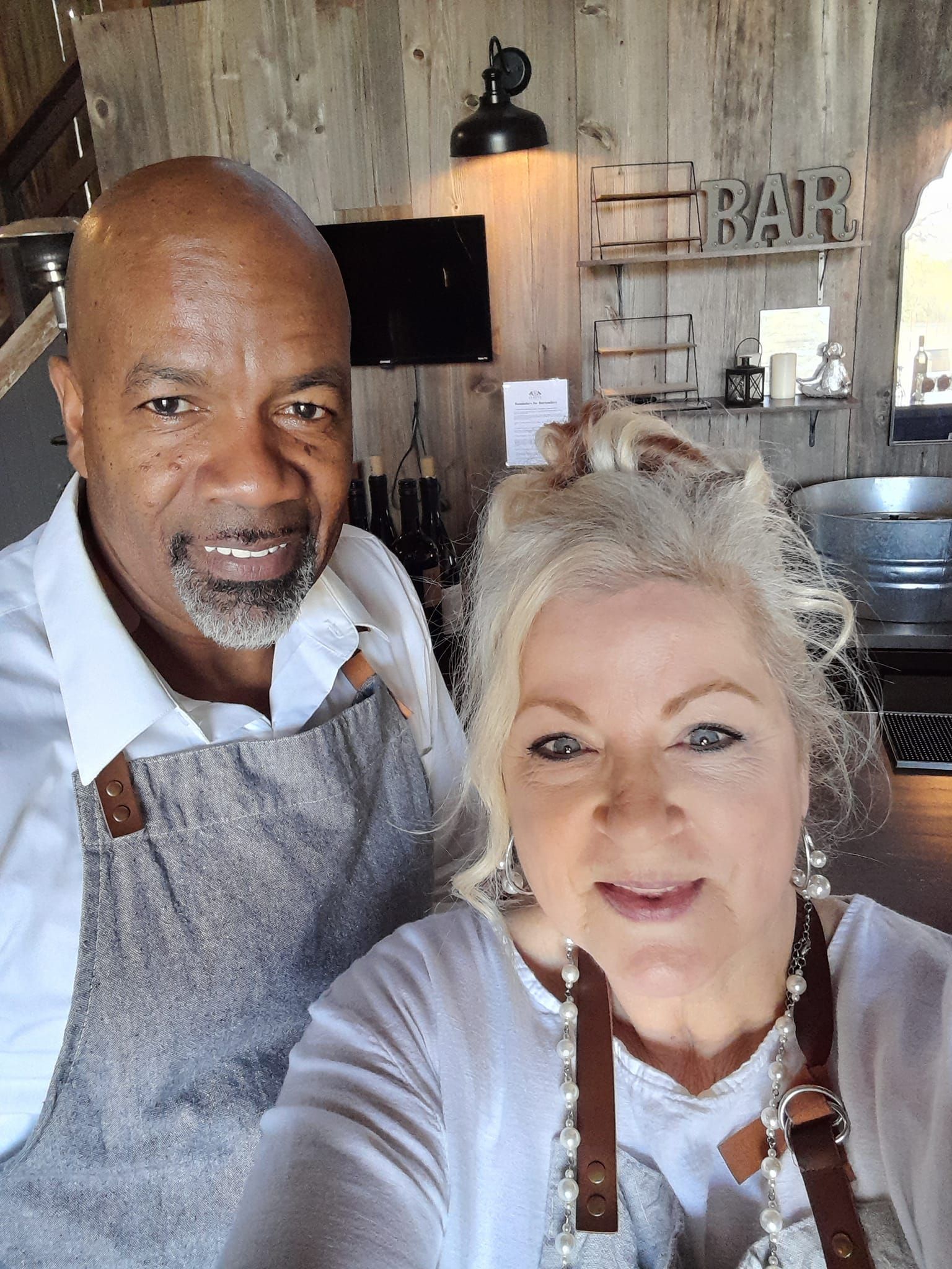 Couple in aprons smile at the camera behind a bar inside a wooden building.